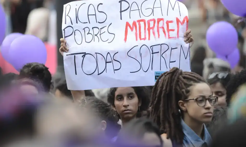 Imagem mostra uma mulher segurando um cartaz escrito: "Ricas pagam, pobres morrem, todas sofrem"