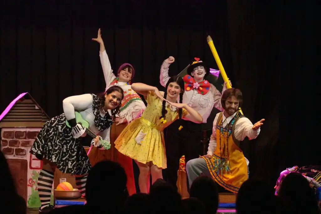 A foto mostra um grupo de atores com roupas coloridas em um palco,