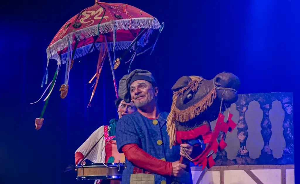 A imagem mostra um homem com roupas coloridas e o rosto pintado, segurando um cavalo de brinquedo.