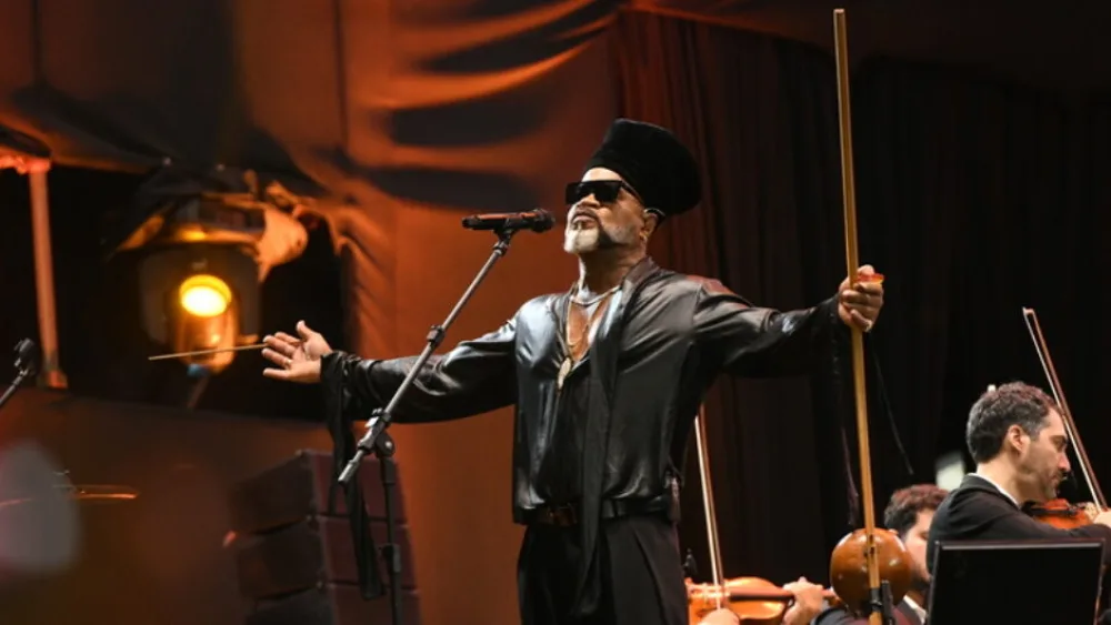 A imagem mostra o cantor Carlinhos Brown vestido de preto e com os braços abertos.