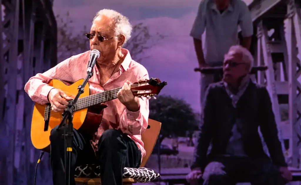 A foto mostra Jards Macalé sentado, tocando violão.
